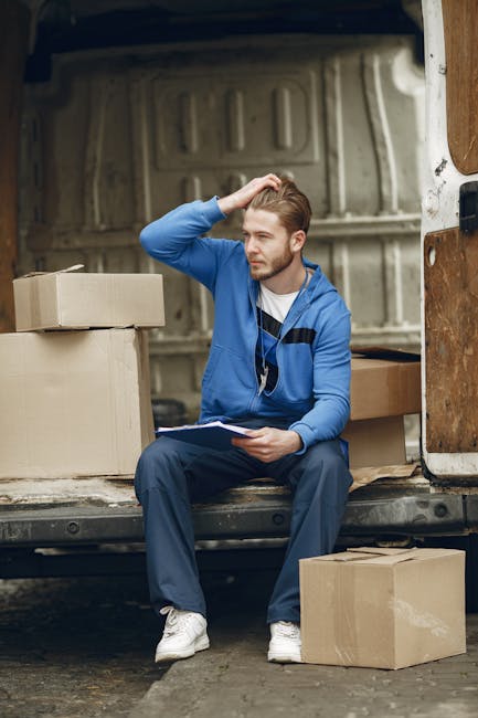 A young man with a beard, wearing a red cap, a brown and beige jacket over a checked shirt, and striped gloves, stands inside the open rear of a van used for house removals. The vehicle's interior sho