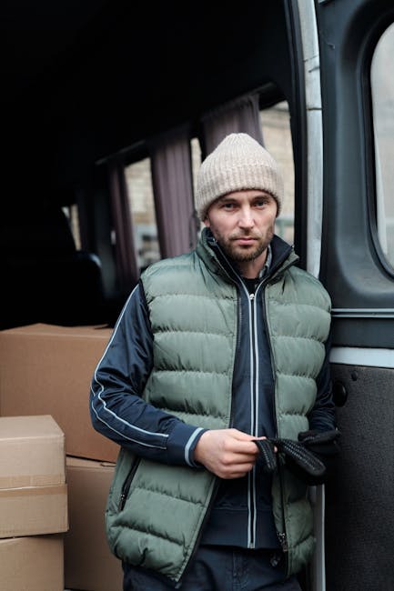 A man with a beard and wearing a black cap, a white polo shirt, and blue jeans is standing in the open rear of a large white van during a home relocation process. He is holding a medium-sized cardboar