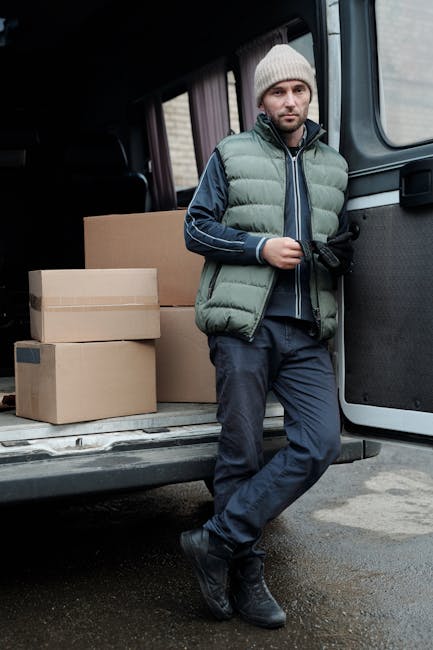 A man dressed in a blue jacket, blue trousers, and white sneakers is loading cardboard boxes into the open rear cargo area of a white van parked on a cobbled street in Haringey. The van, used for home