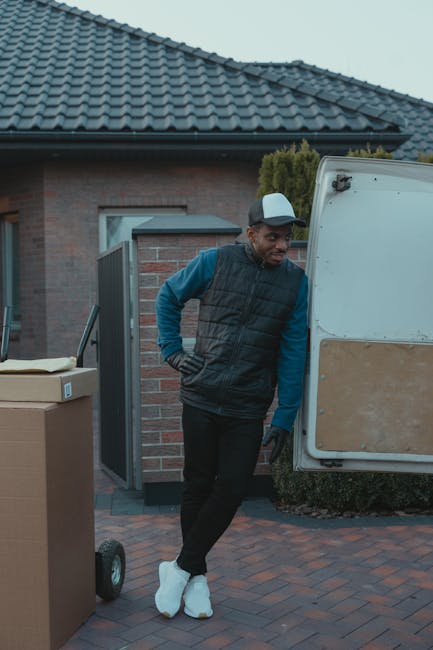 A young man with light brown hair and a beard, dressed in a blue work hoodie with black and grey accents and navy blue work trousers featuring blue pockets, is standing outdoors on a residential stree