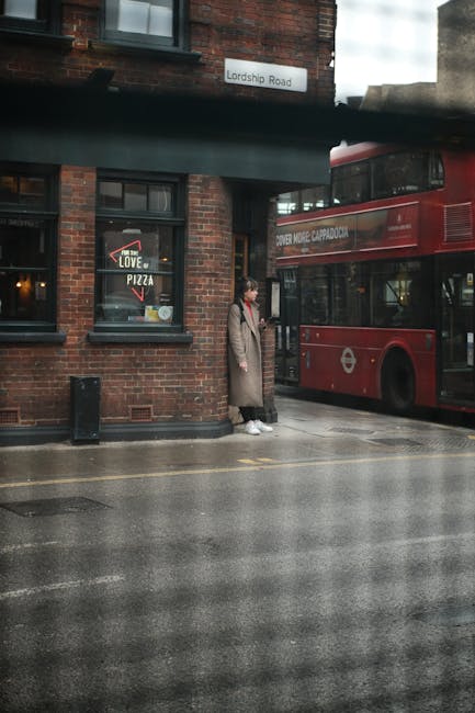 A person standing outside a red double-decker bus on a street in Haringey, on Lordship Road. The individual, dressed in a long beige coat, black trousers, and white sneakers, appears to be waiting or 